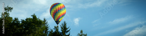 4x1 banner with a flying multi-color balloon against the background of a blue sky with clouds and of a green forest © Anna