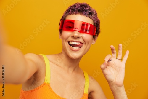 Woman wearing unusual millennial glasses taking selfies in sportswear against an orange studio background, free space