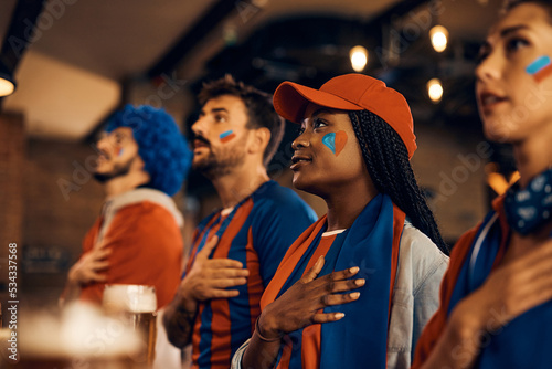 Fotografie Sports fans holding hand on heart and singing national anthem while watching world championship in bar