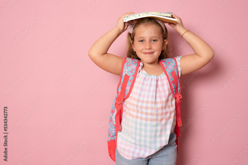 Portrait of a smiling little schoolgirl with backpack holding notebook on head over pink background Portrait of a smiling little schoolgirl with backpack holding notebook on head over pink background