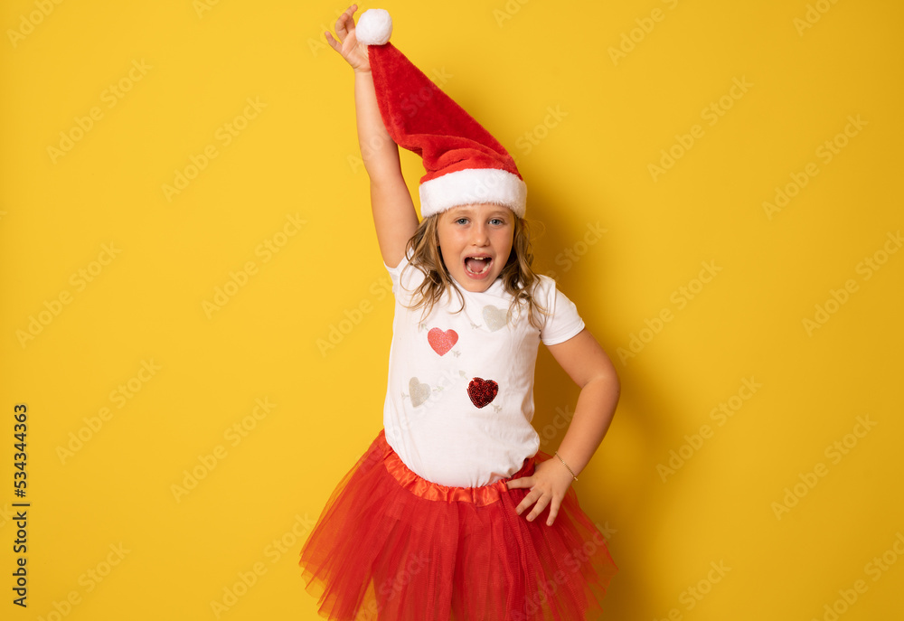 Funny cute little girl in red santa hat standing on yellow background. portrait