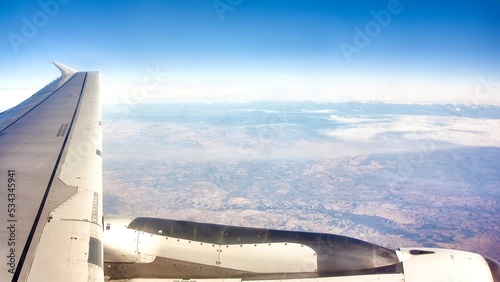 View from the window of an Airbus A321 plane flying above the clouds where we see the engine, a wing and some fields below photo