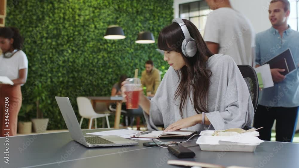 Creative businesswoman, writing and headphones while drinking juice in busy marketing agency, startup and modern office. Productive, focus and young designer listening to music and planning documents Creative businesswoman, writing and headphones while drinking juice in busy marketing agency, startup and modern office. Productive, focus and young designer listening to music and planning documents