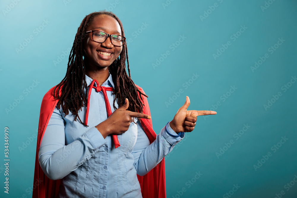 Studio shot of cheerful and optimistic justice defender woman wearing red cape while pointing fingers to right on blue background. Selfless and brave young adult superhero person posing at camera. Studio shot of cheerful and optimistic justice defender woman wearing red cape while pointing fingers to right on blue background. Selfless and brave young adult superhero person posing at camera.