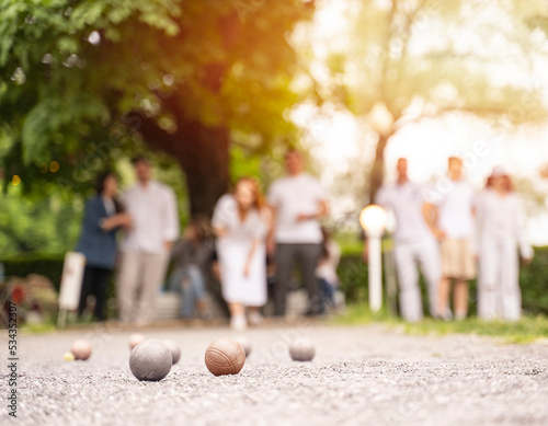 Group of people playing petanque bocce game woman through a ball above green trees park in city park in summer sunset light