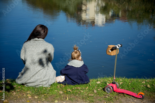 mom and daughter are sitting on the shore of a pond in the park with a scooter