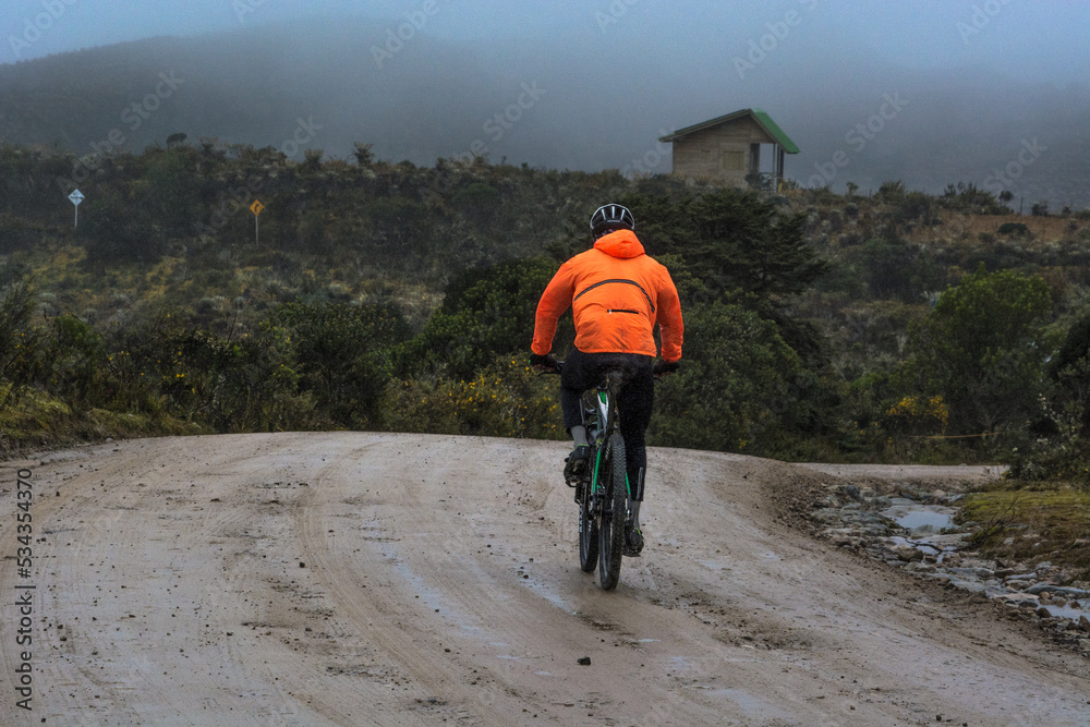 Cyclist man with orange reflective jacket riding mountain bike on a off road path in the mountains. Background with a cabin on the fog and moorland vegetation