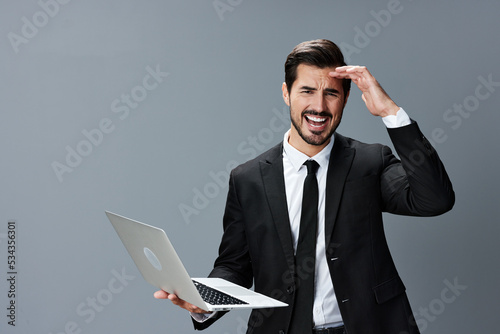 Man with beard brunette business with headache looking at laptop screen on hands on gray background in business suit. Work online business
