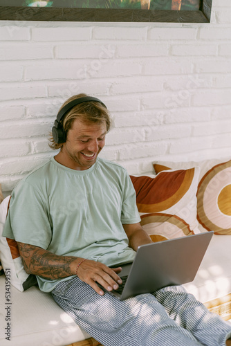 A young smiling man in big headphones works on a laptop in a Bali coworking space. Daily call with the team. Work on your laptop from anywhere.