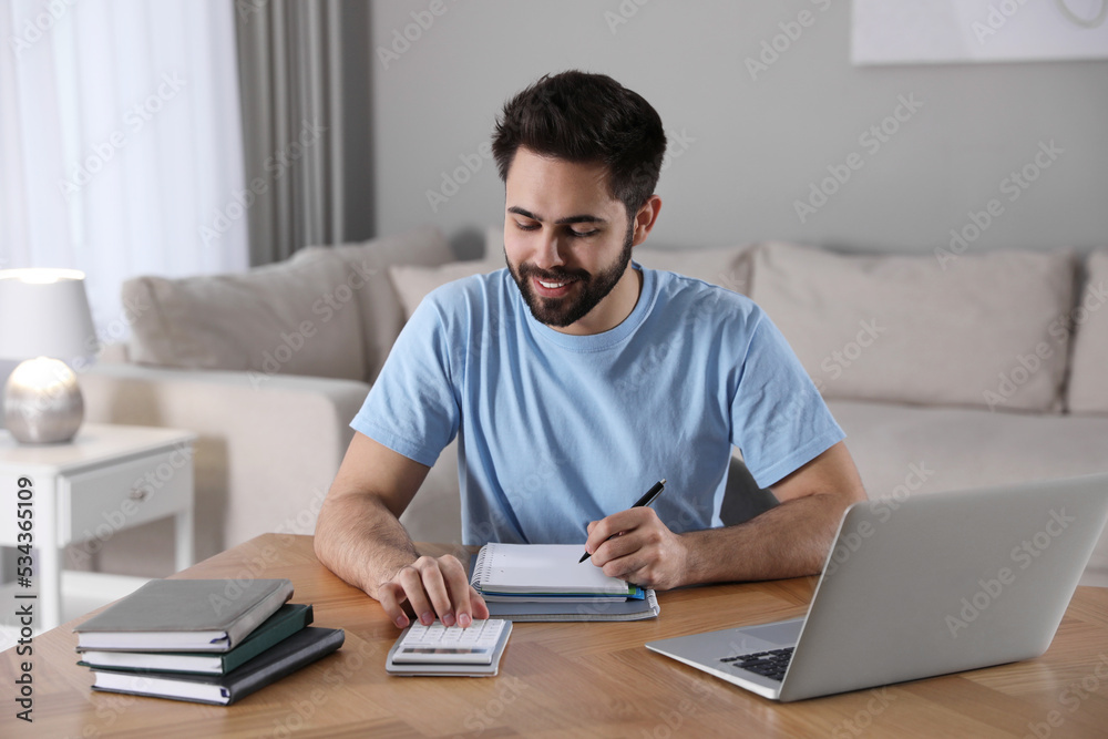 Young man counting on calculator during webinar at table in room Young man counting on calculator during webinar at table in room