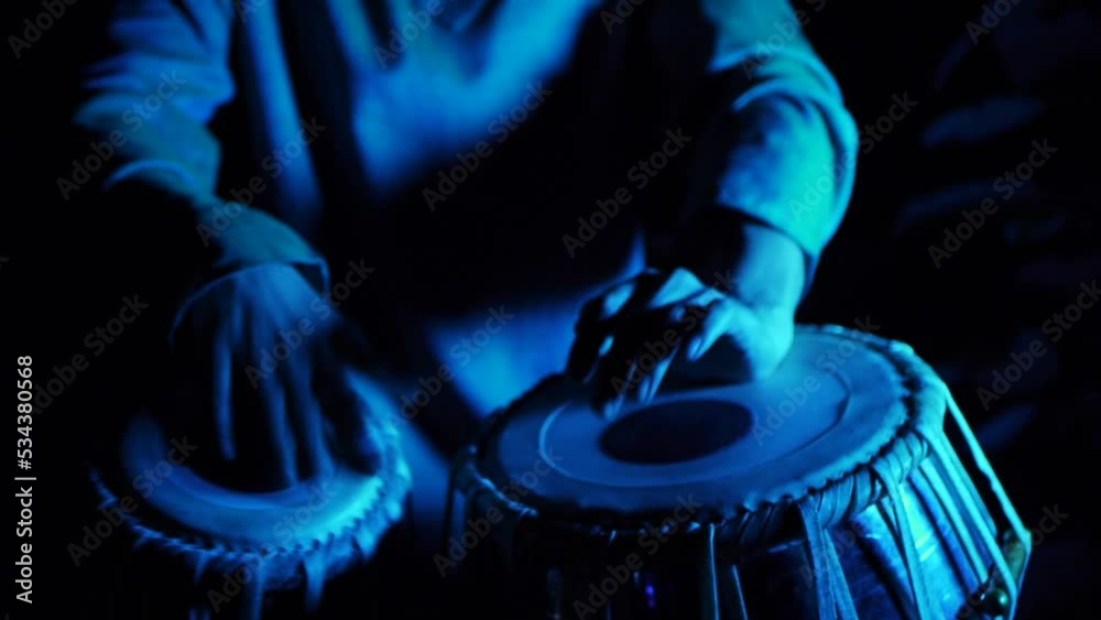 Hands of tabla player drumming intensely on dual tabla setup in dark outdoor area with blue key light, filmed handheld in close up shot