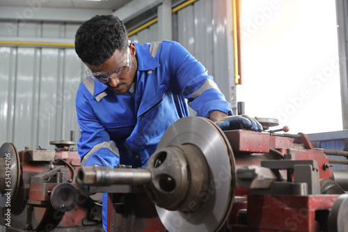 Technician mechanic or worker man in protective uniform using metal lathe machine operate polishing car disc brake at garage. Maintenance automotive and inspecting vehicle part concept