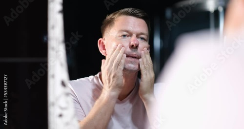 Young man shaves in bathroom and applies aftershave lotion to face photo