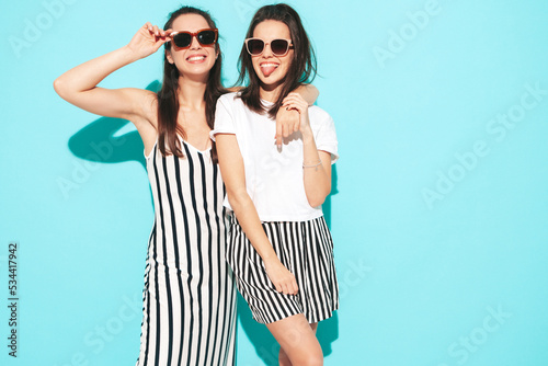 Two young beautiful smiling brunette hipster female in trendy summer shorts clothes. carefree women posing near blue wall. Positive models having fun. Cheerful and happy. In sunglasses