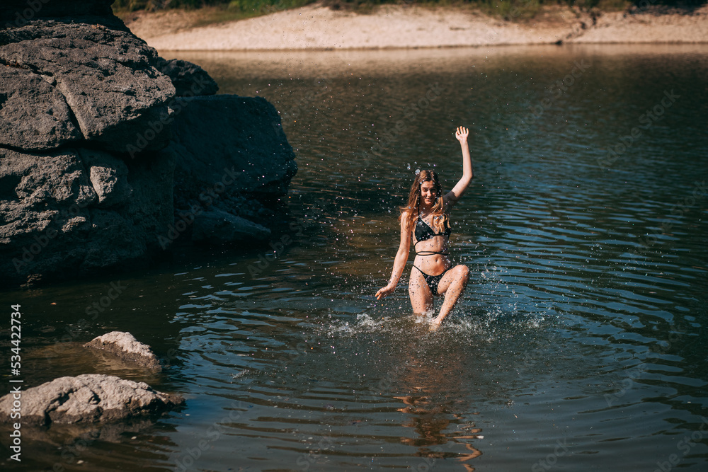 A young girl with long blond hair, a slender figure, in a black swimsuit jumps in the river against the backdrop of rocks and laughs. A young girl with long blond hair, a slender figure, in a black swimsuit jumps in the river against the backdrop of rocks and laughs.