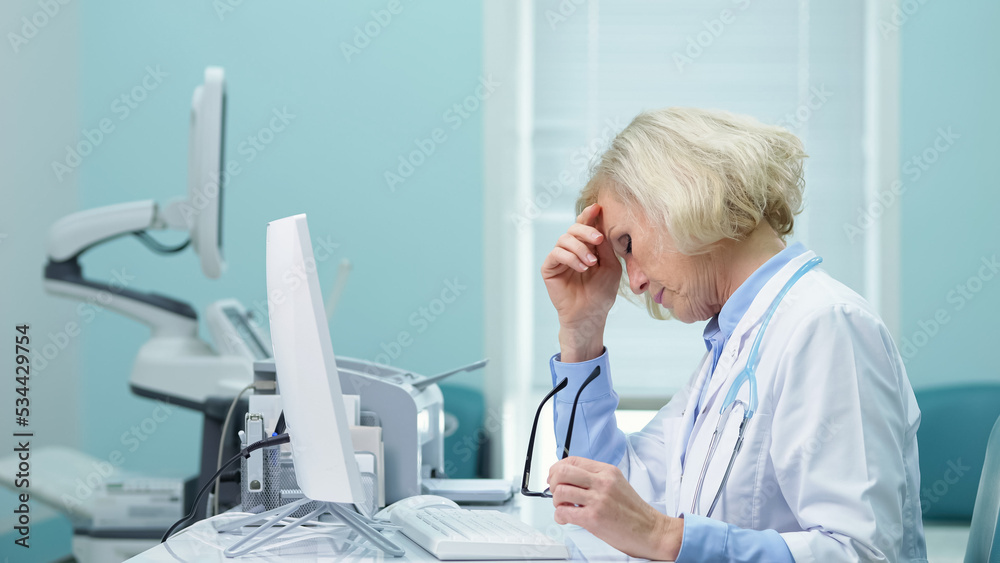 Confident female doctor with glasses enters results of ultrasound diagnostics to main clinic website. Woman eyes hurt from working long at computer monitor