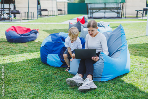 A mother with a laptop, her son sitting next to her outdoors on a green grass lawn. Rest and work with a child photo