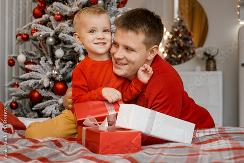 Overjoyed father hugs his charming little son sitting on bed with present boxes near the Christmas tree