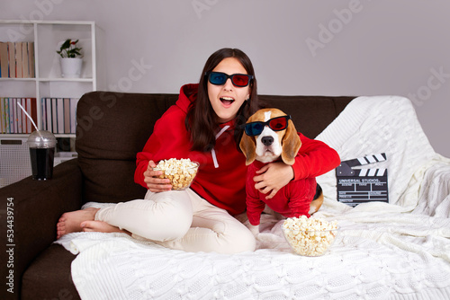 A young girl with her dog beagle in 3 d glasses is sitting on the couch and watching a movie at home.  photo