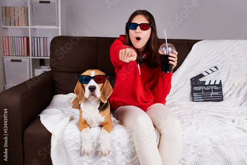 A young girl with her dog beagle in 3 d glasses watching a movie at home. A teenager is holding a glass with a drink in his hands, emotionally pointing his finger at what he saw on TV. photo