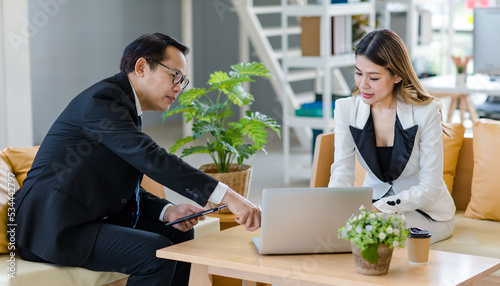 Millennial Asian young successful professional female businesswoman and male businessman colleague in formal suit sitting working with laptop notebook computer together in company office living room
