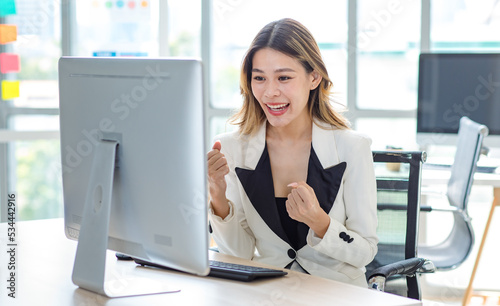Millennial Asian excited successful professional female businesswoman in formal suit sitting smiling screaming holding fists up celebrating victory in front computer screen monitor in company office