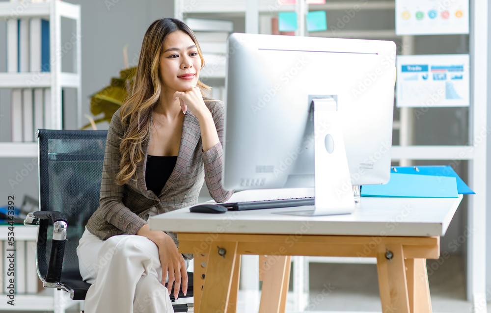 Millennial Asian successful professional female businesswoman employee in formal suit sitting smiling at workstation desk with computer monitor in company office room.