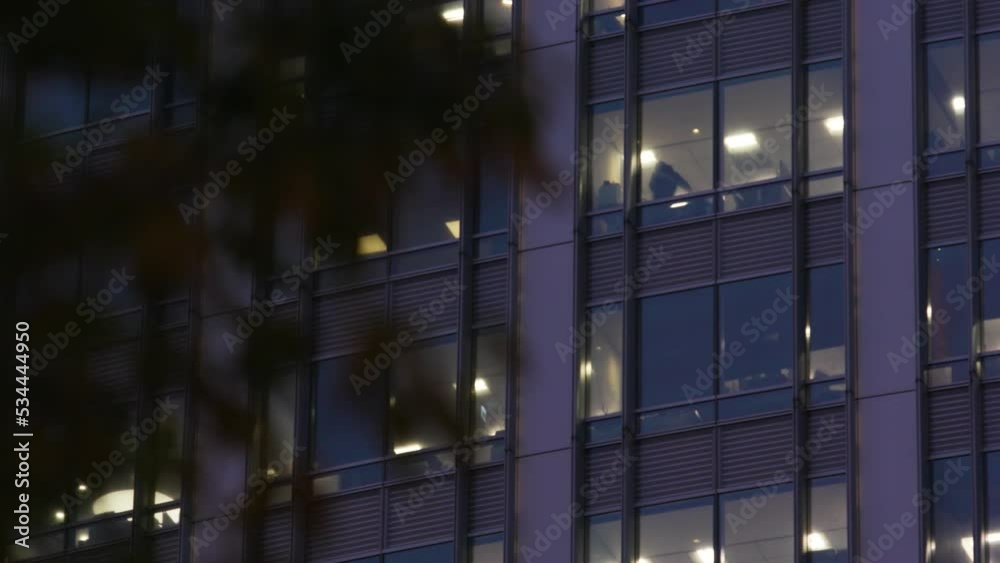 Glass building at night in Taipei with the lights on in the offices inside and silhouettes of employees working late – medium-wide shot. Glass building at night in Taipei with the lights on in the offices inside and silhouettes of employees working late – medium-wide shot.