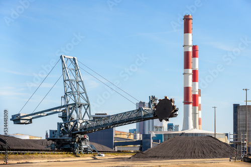 A bucket-wheel stacker-reclaimer and coal stockpiles in a thermal power station with red and white smokestacks on a sunny day.