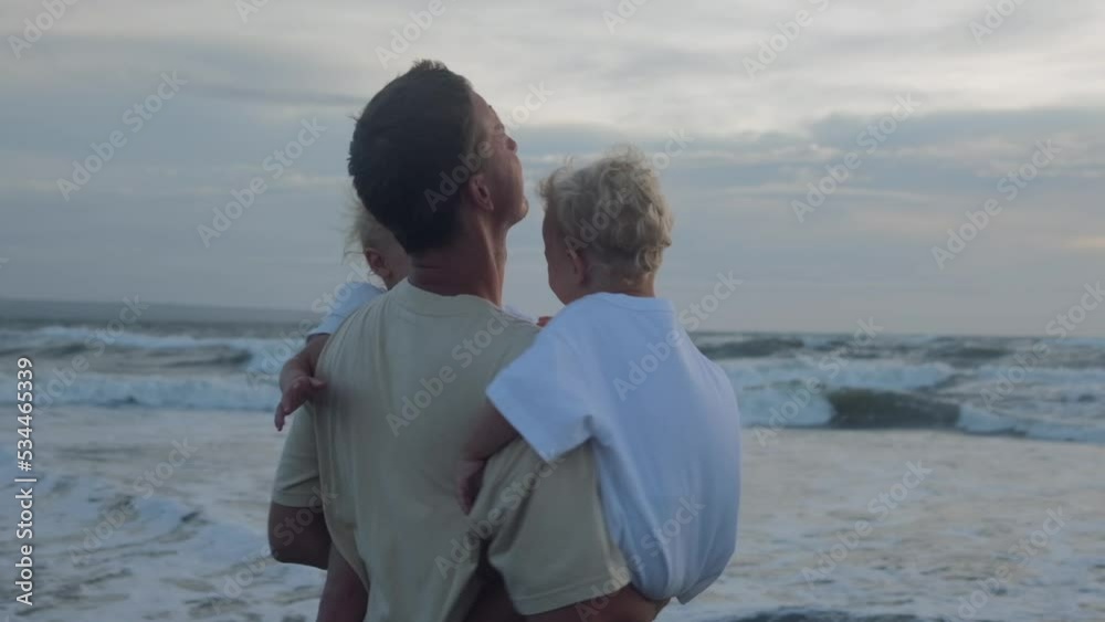 The father of two twin girls holds them both in his strong arms. He circles them, the children are happy and laugh out loud with delight. The seashore is in the background. The father of two twin girls holds them both in his strong arms. He circles them, the children are happy and laugh out loud with delight. The seashore is in the background.