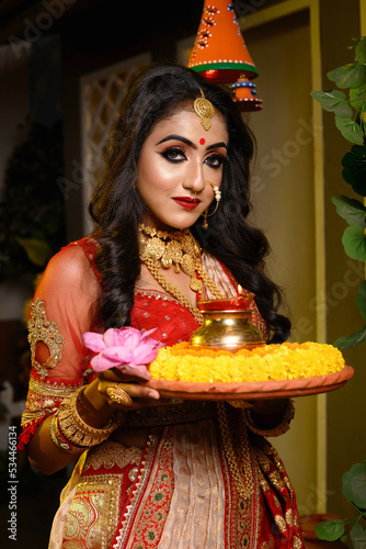 Portrait of pretty young Indian girl wearing traditional saree, gold jewellery and bangles holding plate of religious offering in studio lighting indoor. Indian culture, religion and fashion.