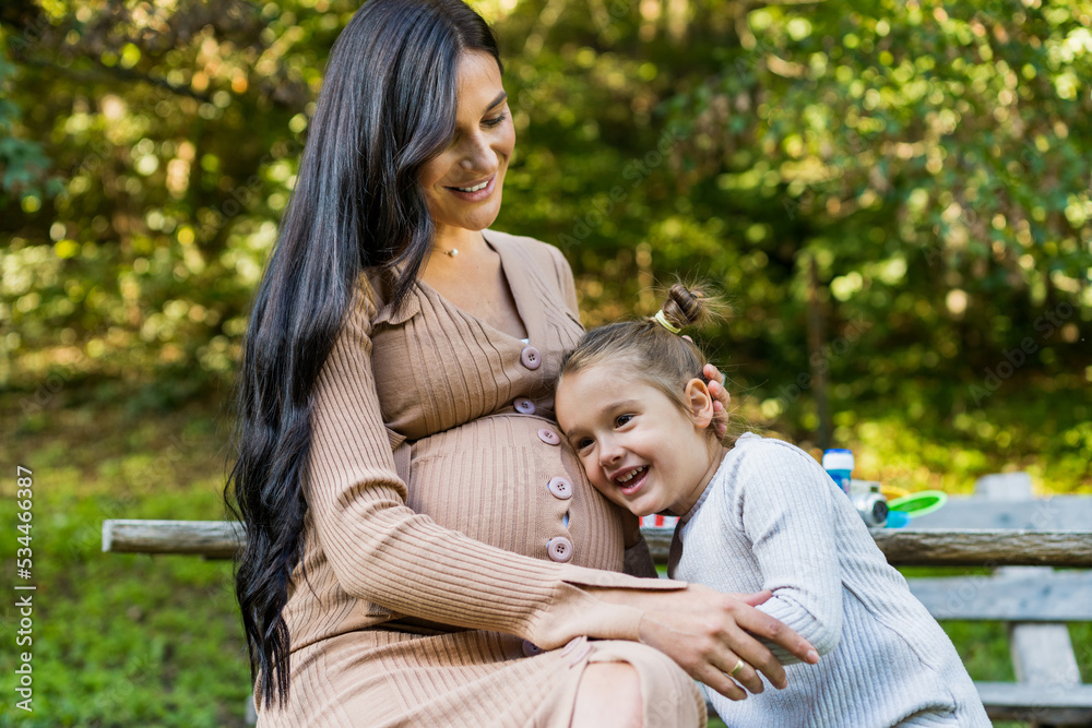 Happy pregnant mother and daughter hugging on a park bench. Happy motherhood. Beautiful woman with a little daughter in nature