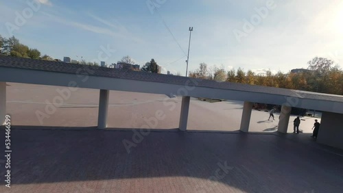 FPV drone view of Guys are skating on the square next to the modern building. Autumn sunny day. Yellow leaves on the trees
