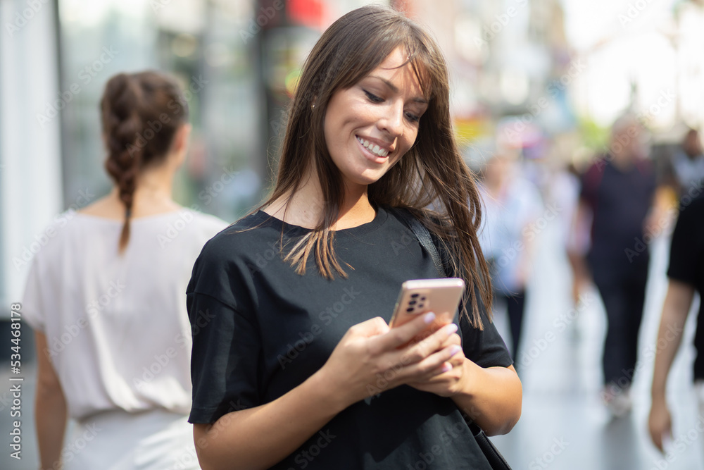 Beautiful smiling woman in black shirt texting on smartphone on street in city center Beautiful smiling woman in black shirt texting on smartphone on street in city center