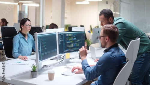 Young male programmer coding on computer looking at screens in office and talking to coworker. Man coding html code and programming on monitor speaking with boss. Web development, developer job