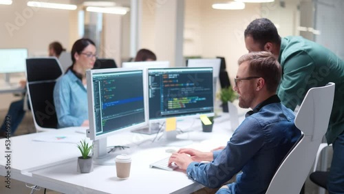 Young professionals writing on sophisticated programming code language and speaking with male boss at office. Team of IT programmers working on desktop computers in data center control room. Workflow