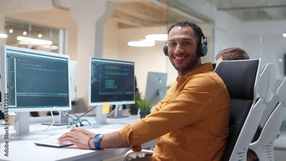 Camera approaching young Hindu man IT programer working on desktop computers typing on keyboard coding and listening to music in headphones. Close up of professional worker in modern office