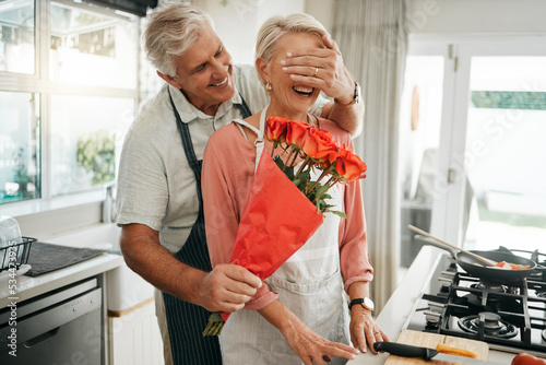 Vászonkép Senior couple, covering eyes and flowers surprise as man give wife bouquet of roses on an anniversary, birthday or valentines day in kitchen