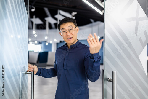 Asian businessman boss invites to his team, man looks at camera and waves, manager in glasses and shirt works inside modern office building.