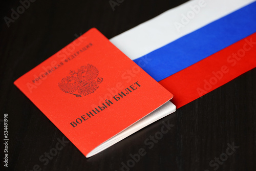 Russian military ID and national flag on wooden table. Translation of inscription: Military ID card, Russian Federation. Concept of mobilization in Russia