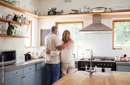 Couple hug  morning coffee and senior people having breakfast in the kitchen while looking at outdoor nature. Elderly man and woman hugging with tea drink in house with love together from behind