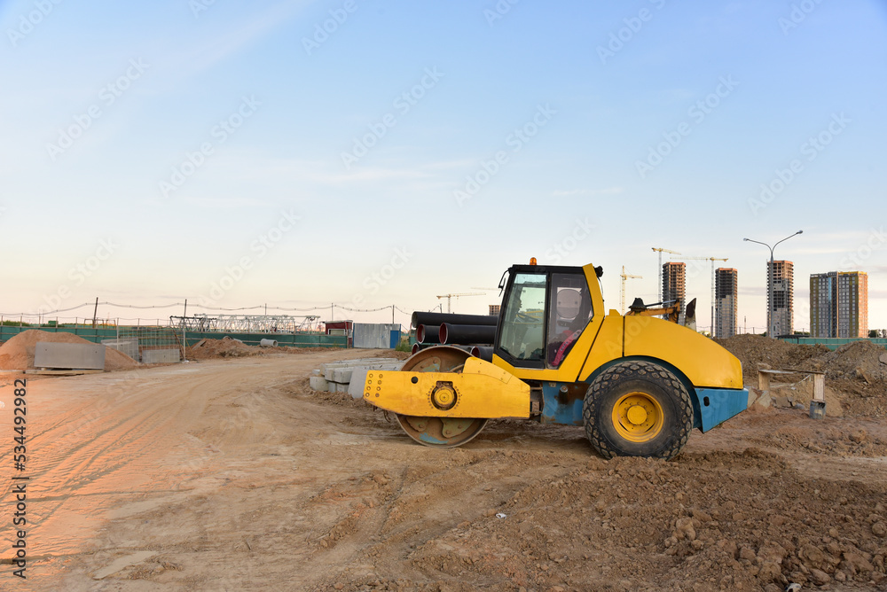 Soil Compactor leveling ground at construction site. Vibration single-cylinder road roller on construction road. Road work and asphalt laying. Tower cranes build high-rise buildings. Road construction