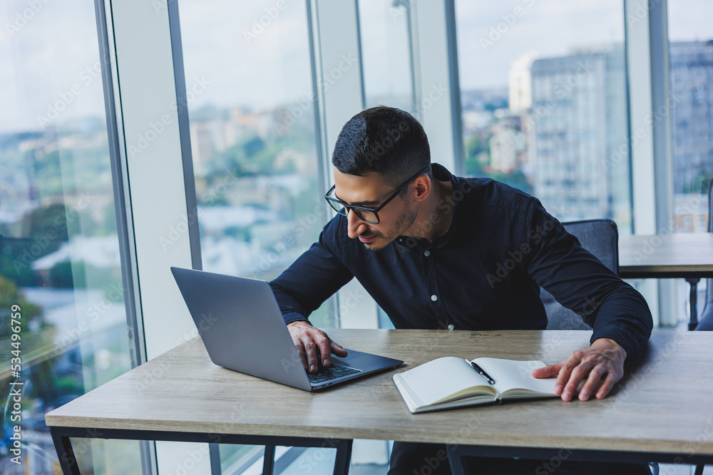 Successful male businessman in glasses and black shirt sits at a desk and works on a project via a laptop in a modern office, looking at the screen. Successful male businessman in glasses and black shirt sits at a desk and works on a project via a laptop in a modern office, looking at the screen.