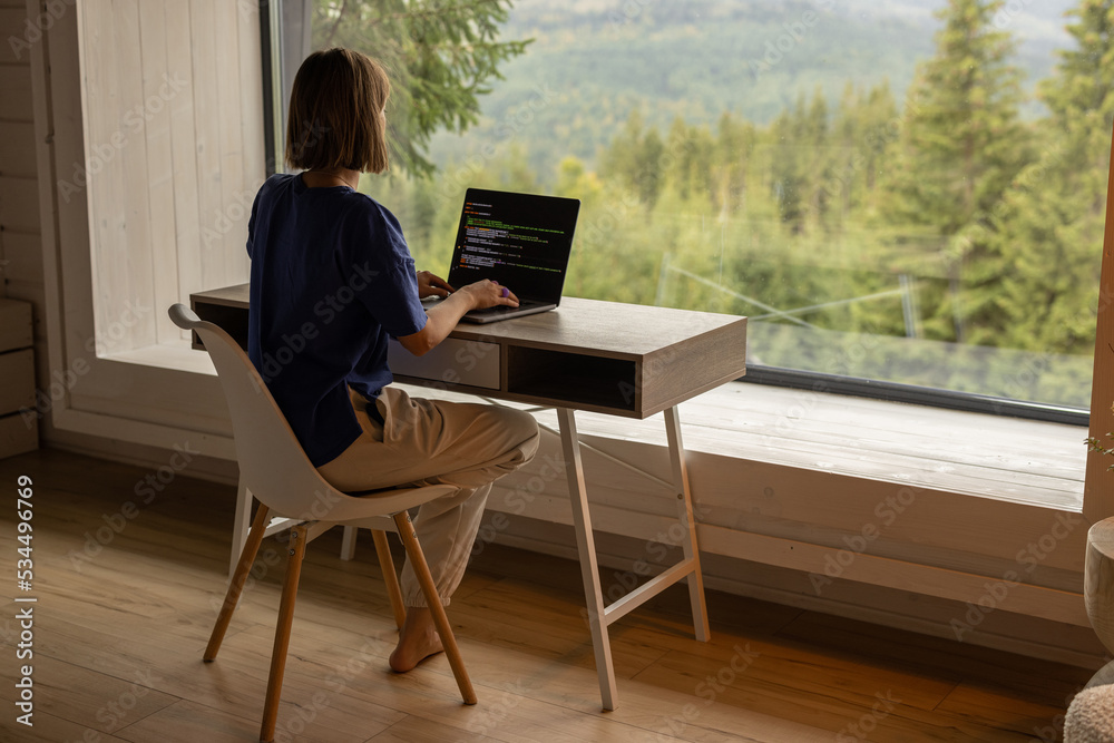 Woman works on laptop while sitting by the table in front of panoramic window with great view on mountains. Remote work and escaping to nature concept Woman works on laptop while sitting by the table in front of panoramic window with great view on mountains. Remote work and escaping to nature concept