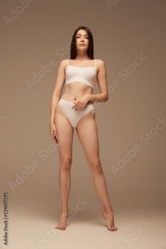 Full-length portrait of young beautiful woman with sportive body posing in white underwear isolated over dark beige studio background