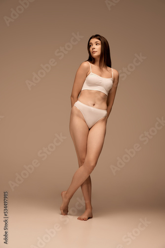 Full-length portrait of young beautiful woman posing in white comfortable underwear isolated over dark beige studio background