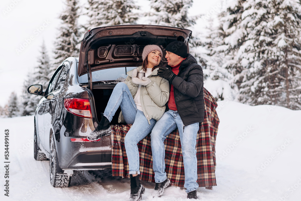 Young couple in love sitting in an embrace in the open trunk of a car,drinking tea in a winter forest.Winter activities,active lifestyle,Valentine's day,tenderness and love.