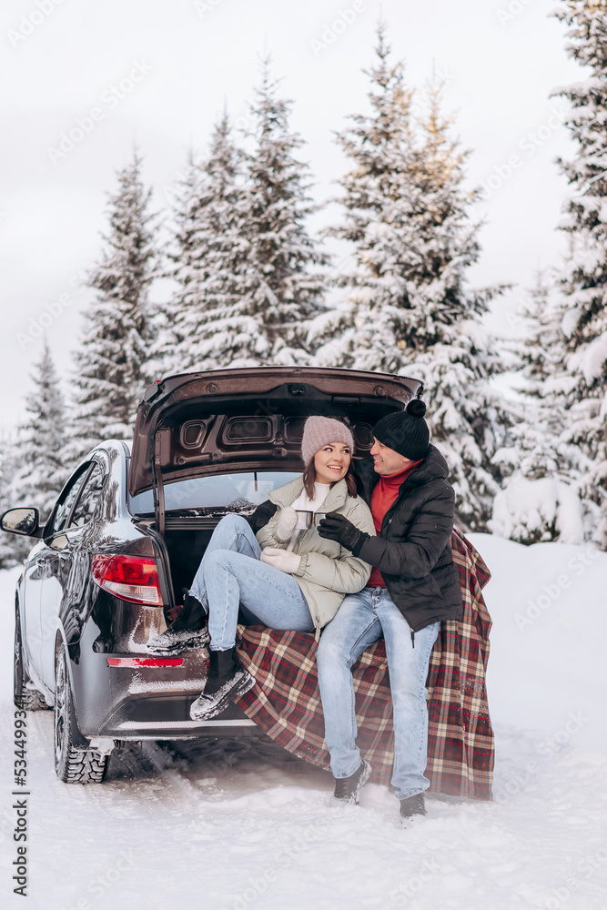 Young couple in love sitting in an embrace in the open trunk of a car,drinking tea in a winter forest.Winter activities,active lifestyle,Valentine's day,tenderness and love.