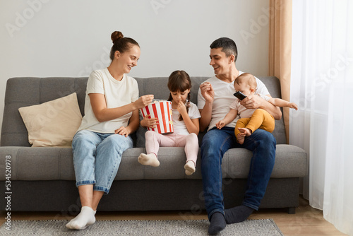 Portrait of loving couple sitting on sofa with their kids and having fun while watching funny video or films, enjoying merry weekend at home, eating popcorn.