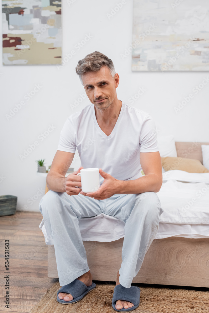 Man in pajamas and slippers holding cup of coffee while sitting on bed.